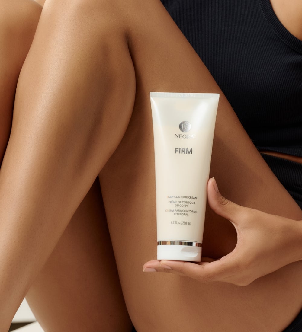 A woman sitting down holding a Firm Body Contour Cream tube in front of her legs.